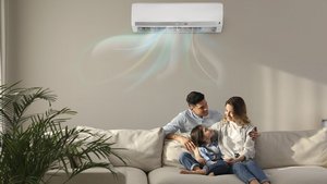 Im Wohnzimmer sitzt eine Frau mit Tochter auf dem Sofa und der Mann steht hinter den beiden. Alle schauen sich glücklich an. An der Wand im Hintergrund ist eine Klimaanlage, die Frische Luft in den Raum bringt.